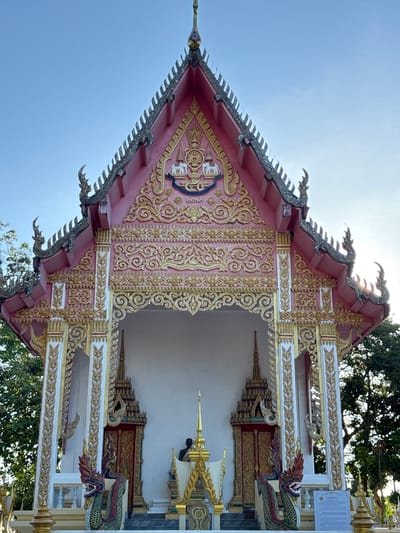 A low-angle, full shot captures the ornate facade of a Buddhist temple in Baan Tha Po, Thailand. The temple's roof is a vibrant pink, trimmed with gold and intricate architectural details. Decorative, mythical serpent-like figures adorn the roofline. The entrance features a grand archway with detailed golden carvings and two towering white pillars, also decorated with gold patterns. Flanking the entrance are smaller, red-doored structures topped with golden spires and similar serpent motifs. In the foreground, two prominent, colorful naga (serpent) statues with fierce expressions guard the entrance. Behind the naga, a dark-robed figure is visible inside the temple's entrance. Several golden spires rise from the temple grounds. Green trees and a clear blue sky form the backdrop. A white sign with Thai writing is visible on the right side of the image.