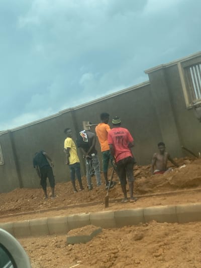 The image captures a group of approximately six men gathered on a dirt-covered worksite next to a concrete building, under an overcast sky. The photograph is taken from a slightly elevated perspective, looking down at the subjects and the ground.
The scene depicts an outdoor setting, likely during daytime, characterized by a predominantly brown, uneven dirt ground. A light-colored concrete curb or pathway runs vertically through the middle-right of the frame, separating different sections of the ground.
The men are spaced along the edge of the building on the left. From right to left: one man is shirtless, wearing red shorts, standing on a higher earthen bank. Next to him, another man wears a red t-shirt with the word "GADA" printed in white on its back, possibly holding a tool that extends downwards. Following him is a man in an orange t-shirt, and then another in a dark hoodie with a colorful graphic. A fifth man is seen in a bright yellow t-shirt and dark trousers, facing towards the left. The man furthest to the left is dressed in a dark t-shirt and dark trousers, with his back partially turned.
The building is a concrete structure with visible architectural details like horizontal slats on its upper levels. The sky is a pale, cloudy blue, suggesting an overcast day. The overall impression is one of laborers or workers at a construction or development site in Gosa, Nigeria.