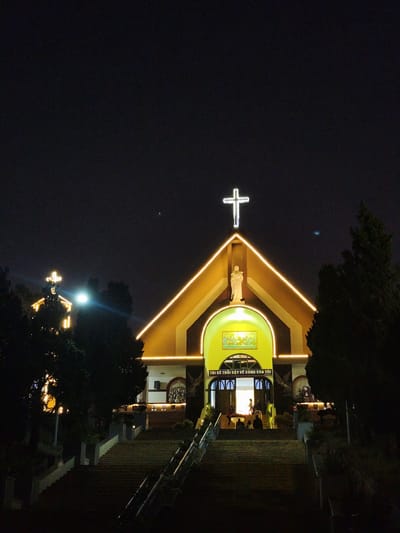 The image displays the Domaine de Marie Church in Dalat, Vietnam, captured at night. The central focus is the main church building, characterized by a prominent gabled roof outlined with warm yellow string lights. A large, bright white cross stands atop the roof's apex. Below the cross, a white statue of a robed figure, likely the Virgin Mary, is positioned in a central niche above the main entrance. The facade of the church is rendered in shades of warm yellow and orange, illuminated by internal and external lighting.
Below the statue, an arched entryway glows brightly from within, revealing the interior space. Above the arch, Vietnamese text reads "TÔI SẼ TRỞI DẬY VỀ CÙNG CHA TÔI" (I WILL RISE TO MY FATHER). Further above this inscription, a rectangular mosaic or painting depicting multiple figures is visible.
A wide, multi-tiered concrete staircase with handrails leads directly from the foreground to the church's main entrance. Several individuals are present on these steps and within the illuminated entrance. Some figures are seated on the lower steps, while others are standing or walking near the entrance, appearing to gather or exit. Their actions are indistinct due to distance and lighting.
To the left of the main church, a smaller, similarly gabled building is visible, also illuminated with warm yellow lights and topped with a smaller white cross. This building is partially obscured by dark evergreen trees. Additional dark evergreen trees flank the staircase and the church buildings, contrasting sharply with the illuminated structures. The sky is dark, indicative of nighttime, with a subtle light source visible in the upper left background.