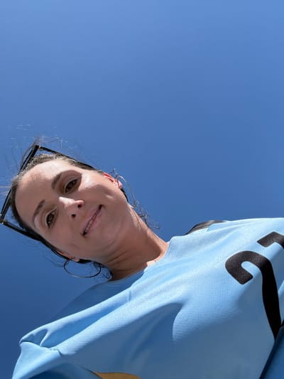 This is a low-angle, first-person selfie of a woman against a bright blue sky. The woman has dark hair, brown eyes, and is smiling. She is wearing a light blue t-shirt with black lettering on the front. She is also wearing large hoop earrings and sunglasses are pushed up on her head. The photo appears to have been taken outdoors on a clear, sunny day. No specific location in Miami Gardens is discernible from the image itself.