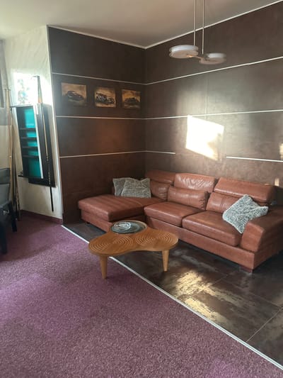 The image depicts an indoor scene, likely a living room or lounge area. A large, brown leather sectional sofa with plush cushions is the central furniture item. In front of the sofa, a unique, wooden coffee table with a distinctive swirl pattern rests on a dark tiled floor. To the left, a portion of a dark-colored carpet is visible, transitioning to the tiled floor. On the wall behind the sofa, three framed artworks featuring vintage cars are displayed. To the far left, a tall, dark shelving unit contains items and billiard cues stand vertically beside it. A modern ceiling light fixture with two circular shades hangs from the ceiling. Sunlight streams into the room, creating bright reflections on the wall and floor. The overall ambiance suggests a comfortable and somewhat eclectic living space. The location is identified as Brca, Montenegro.