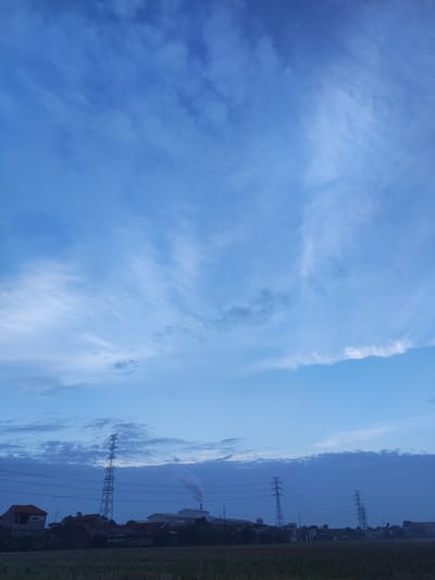 The image depicts a rural or semi-rural landscape in Banjarkemantren, Indonesia, under a vast, cloudy blue sky, likely during dusk or dawn. In the foreground, a field of dry grass or crops stretches horizontally. The middle ground reveals a collection of low-rise buildings, including houses with terracotta roofs and industrial structures. Prominently featured are several tall electricity pylons and power lines crisscrossing the scene. One industrial building emits a plume of smoke from its chimney, suggesting ongoing operations. The overall atmosphere is calm and serene, with the muted light casting long shadows and softening the details of the landscape.