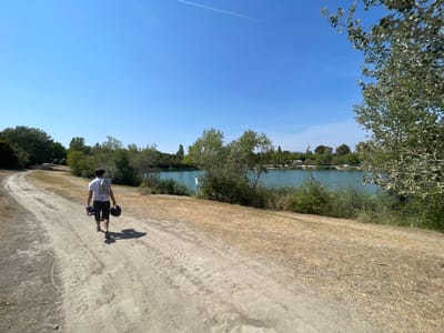 A person walks on a dirt path along the edge of a body of water on a clear, sunny day. The path is dusty and has tire tracks, suggesting it is used by vehicles. To the right of the path, dry, yellow grass covers the ground. Beyond the grass is a dense line of green bushes and trees that leads to a bright turquoise body of water. The water appears calm with a few ripples. In the distance, across the water, is a shoreline with more trees and some buildings. The sky is a vibrant, clear blue with a faint contrail from an airplane. The person is wearing a white t-shirt, dark shorts, and sneakers. They are carrying a black helmet and a dark object, possibly a skateboard or bag, in their left hand, and a grey towel or shirt draped over their left shoulder. The person is walking away from the camera, towards the body of water. The scene is likely in a park or recreational area near a lake or reservoir. This location is situated in Cadenet, France, given the context provided.