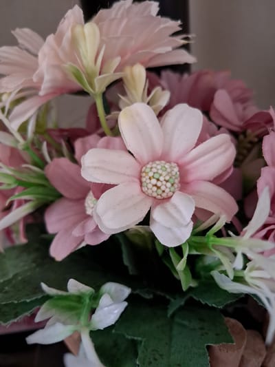 This close-up image showcases a bouquet of artificial flowers, likely part of a decorative arrangement, in Gavar, Armenia.
The central and most prominent flower features soft, light pink fabric petals with a distinct, beaded center composed of small, spherical elements in alternating light green and white. Surrounding this main bloom are other artificial flowers in various shades of pink, from pale to a deeper rose, and creamy white. Some smaller, unopened buds are also visible, hinting at different stages of bloom.
Interspersed throughout the arrangement is artificial green foliage, including leaves and stems, which provide a natural contrast to the floral elements. The texture of the fabric petals and leaves is visible, indicating their artificial nature. In the background, particularly towards the right side of the image, there are glimpses of a brown, rough-textured material, possibly cork or another decorative filler, adding depth to the arrangement.
The lighting is soft, highlighting the colors and textures of the flowers. The depth of field is shallow, bringing the artificial blossoms into sharp focus while gently blurring the background. No activity is taking place, and no people or visible text are present in the image. The scene suggests an indoor setting or a shaded outdoor area where the decorative flowers are displayed.