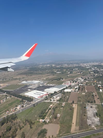 An aerial view from an airplane window captures a diverse landscape in Santa María Zacatepec, Mexico, likely during daytime under a clear blue sky. The left side of the frame features the white and red wingtip of the aircraft, with a portion of the wing and flaps visible, indicating the plane is in flight, possibly during ascent or descent.
The ground below presents a mix of urban development, industrial areas, and agricultural fields. In the foreground and midground, there are numerous buildings, including large white warehouses or factory structures with extensive parking lots filled with vehicles. Adjacent to these industrial areas, residential buildings and smaller structures are interspersed with green patches of trees and cultivated fields, some appearing barren or recently tilled. A prominent road or highway stretches across the scene, with cars visible, suggesting active transportation. Further in the distance, a sprawling town or city extends into the hazy horizon, bordered by what appear to be mountains or elevated terrain. The overall impression is one of a developing suburban or industrial region transitioning into more rural areas. No discernible text is visible on any buildings or signs. The weather appears clear with good visibility, although a slight atmospheric haze softens the distant landscape.