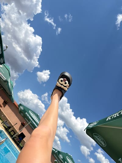 The image shows a person's leg and foot, wearing a black sandal with a cartoonish face design, extended upwards against a bright blue sky filled with white, fluffy clouds. The background includes green umbrellas, a building with a terracotta-colored roof, lounge chairs, and a swimming pool. The scene appears to be an outdoor recreational area, possibly a resort or a public pool, under sunny conditions. The overall impression is of leisure and relaxation.