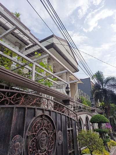 A large ornate black and copper gate stands before a multi-story house with a pergola-like structure adorned with greenery. The house features white columns, a brown roof, and a balcony. Lush landscaping includes shaped trees and palms. Overhead, several power lines crisscross the sky against a backdrop of scattered clouds.