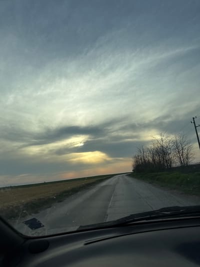 The image shows a view from inside a car, looking out onto a straight, paved road. The road appears to be wet. To the right of the road is a field. A line of bare trees is visible on the right side of the road. The sky is cloudy with patches of sunlight visible. The location is likely rural, potentially in or near Sofronievo, Bulgaria, given the mention of the city. There are no people visible in the image. A power line and pole are visible in the distance.