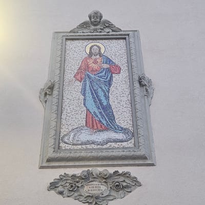 The image shows a mosaic depicting Jesus Christ with a radiant heart, set within an ornate frame. Above the mosaic, a sculpted cherub with outstretched wings is visible. Below the mosaic, a decorative plaque with floral motifs features an inscription that reads: "INDULGENZA DI 50 GIORNI A CHI RECITA UN GLORIA PATRI MONS. VESCOVO M. 4 MARZO 1925". This inscription indicates an indulgence of 50 days for those who recite the Gloria Patri, granted by a bishop on March 4, 1925. The artwork is mounted on a textured, light-colored wall, likely an exterior building facade, in Calcinate, Italy. The overall style suggests religious or devotional art.