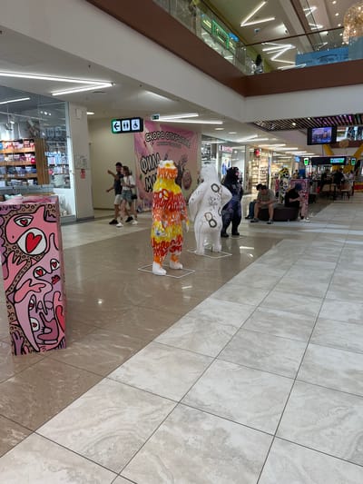 The image depicts the interior of a modern shopping mall, likely in Yazvitsevo, Russia, given the context. In the foreground, a colorful, abstract art installation of a figure resembling a bear stands on a white base. To its left, another art installation, a pink, rectangular prism covered in abstract faces and hearts, is also visible.
Further back, several people are walking and standing. Two individuals, a man and a woman, are seen in conversation. In the middle ground, three more art installations of bear-like figures, one white with black markings, another black with purple designs, and a third with a yellow and orange fiery pattern, are placed. Several shoppers are seated on benches, some looking at their phones.
The environment is characterized by polished tile flooring, bright overhead lighting, and store displays visible in the background. Signage indicating "Coffee Like" suggests a food and beverage outlet. A banner with Cyrillic text, reading "СКОРО ОТКРЫТИЕ" (Soon Opening), is prominently displayed. Signs for restrooms and accessibility are also visible on a hanging panel. The overall scene conveys a public, commercial space with an artistic installation as a focal point.
