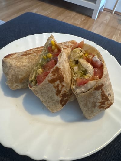 The image shows a white plate with three sections of a sliced burrito. The burrito appears to contain chicken, corn, tomato, and a green sauce, likely guacamole or a similar avocado-based dressing. The tortilla is a brown, speckled variety. The plate sits on a blue surface, possibly a placemat or tablecloth. In the background, a glimpse of wooden flooring and a white table leg are visible. The location is not apparent from the image, thus I cannot include Subotica, Serbia, in my description.