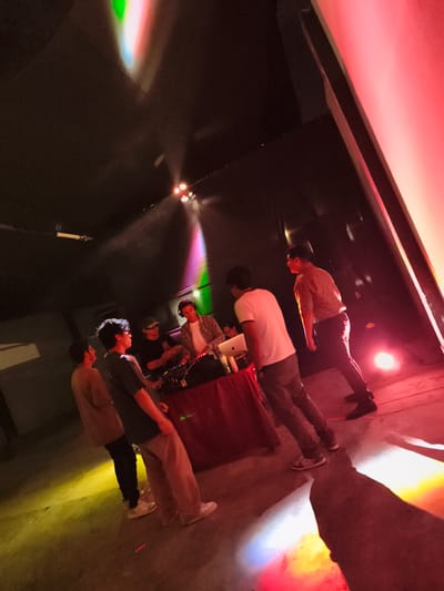 The image captures a dimly lit scene, likely indoors, possibly in Chimbote, Peru. Several people are present, appearing to be at a small gathering or performance. A person at a table is using a mixing console and laptop, suggesting a DJ or musical performance. Attendees are standing around. The lighting is colorful, with beams of green, red, and yellow illuminating the space. The floor and walls are bare.