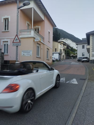 The image depicts a street scene in Orselina, Switzerland, likely on a cloudy day. A white convertible is captured in motion in the foreground, its front left wheel and a portion of its side visible. The car appears to be a Volkswagen Beetle. On the right side of the street, a dark SUV is parked. Further down the street, another dark SUV is visible. In the background, a multi-story building with a pale pink facade is prominent, featuring several windows and a balcony. A street sign indicating "SCUOLE" (schools) with a pedestrian crossing symbol is mounted on a pole near the pink building. The road surface is asphalt, marked with white directional arrows. A steep, tree-covered hill rises in the background, with a few buildings visible on its slopes. The overall impression is a residential or mixed-use area with traffic and pedestrian infrastructure.