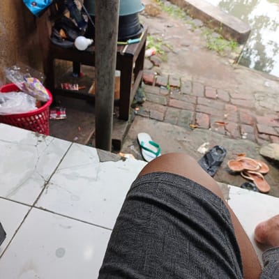 The image is taken from the perspective of a person sitting on a white tiled floor, looking towards a brick pathway. Their right leg is visible, clad in grey patterned shorts. The background features a partially obscured wooden table with a white light bulb hanging above. A red plastic basket with loose objects inside sits on the left side of the frame. Other items, like a plastic water jug and a blue bag, are faintly visible in the upper left corner. The environment is a weathered outdoor area with a brick path and a small body of water in the background. The location is likely in a residential area in Pameungpeuk, Indonesia.