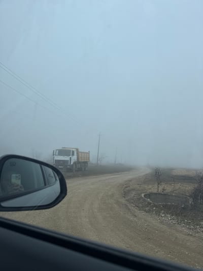 The image depicts a rural scene characterized by dense fog, located in Зеленогорское сельское поселение, Ukraine. The perspective is from the interior of a vehicle, looking out the passenger-side window and into the side mirror. A heavy, uniform light grey fog significantly reduces visibility across the landscape.
In the midground, a white dump truck with a light brown bed is stationary on the left side of an unpaved, dirt road. The truck is oriented towards the viewer's vehicle. The dirt road itself, showing visible tire tracks, curves right and recedes into the fog. To the left of the road, multiple wooden utility poles support overhead power lines, which stretch horizontally across the frame before disappearing into the mist. Sparse, leafless trees and shrubs are visible along the roadside and in the surrounding fields, indicative of a dormant season.
In the immediate foreground, the dark dashboard and door trim of the viewer's vehicle are visible. The passenger-side mirror prominently reflects a person holding a smartphone, actively capturing the scene. To the right of the dirt road, a partially visible circular concrete structure, resembling a well casing or culvert, is present on the dry, uneven ground. The overall environment is one of limited visibility and a muted, cool color palette.