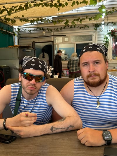 Two men, dressed in blue and white striped tank tops and black bandanas, sit at a wooden table. The man on the left wears mirrored sunglasses and a ring with a pink stone. The man on the right has a beard and wears a necklace with a tooth-shaped pendant. Behind them, several other people are visible in the background. The setting appears to be an outdoor cafe or restaurant with a canopy overhead, decorated with hanging vines and string lights. The scene is likely in a casual, open-air establishment.