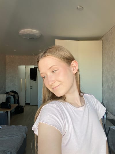 A young woman with long, straight light brown hair and light-colored eyes is captured in a close-up, horizontal portrait, smiling softly. She is wearing a light pink, short-sleeved t-shirt with a ruffled or lettuce-edge trim on the collar and sleeves. Her head is slightly tilted, and her gaze is directed upwards and to the viewer's left, conveying a pleasant and relaxed emotion. Her skin has a fair complexion, and she appears to be wearing minimal makeup, with a hint of blush on her cheeks.
The setting is an indoor room, likely a bedroom or living area, located in Зубово, Russia. The woman appears to be reclining or lying down, with the background objects positioned above and around her. To her left, a light gray wall features a circular recessed light fixture and a white, oval-shaped surface-mounted device, possibly a smart home sensor or smoke detector. Above her, a light-colored, textured ceiling is visible. To her right, a wardrobe or cabinet with light-colored doors stands in the background. Further back and below the wardrobe, shelves or drawers are visible, holding items such as black fabric and small containers. A dark blue or gray upholstered furniture piece, possibly a sofa or bed, covered with a light-colored throw blanket, is also present in the background. The room is brightly and evenly lit, suggesting either natural daylight or well-diffused artificial lighting. There is no visible text in the image.
