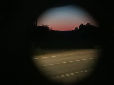 The image shows a dark road with white lane markings at dusk, viewed through an oval-shaped aperture, suggesting it was taken through a lens or opening. Silhouetted trees and a dark horizon are visible against a twilight sky with hues of pink, orange, and blue. The road appears wet and illuminated by artificial light, possibly headlights. The location is specified as Воскресенское, Russia.
