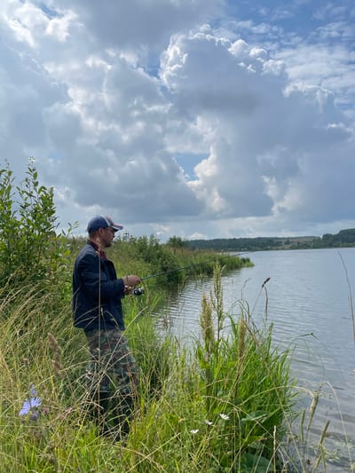 A man is fishing on the bank of a body of water, possibly a river or lake, in Bereznik, Russia. He is wearing a dark jacket, camouflage pants, and a baseball cap. He holds a fishing rod with a reel in his right hand. The surrounding vegetation is tall grass and reeds. The sky is partly cloudy with dramatic cloud formations. The water is calm, reflecting the sky. In the background, there is a wooded shoreline and hills. The overall scene depicts a serene outdoor activity in a natural environment.