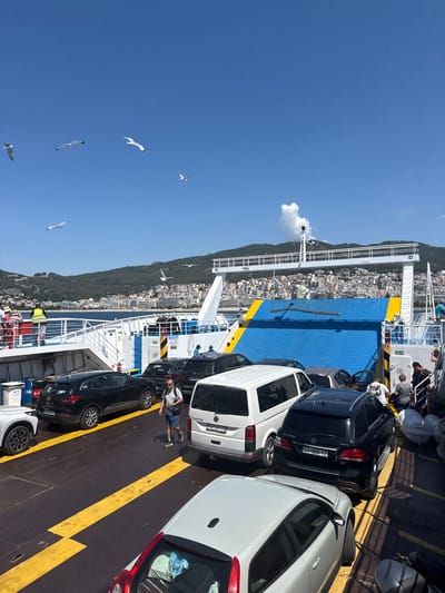 This image depicts a ferry carrying vehicles and passengers, likely in the port of Kavala, Greece, given the cityscape and hilly terrain visible in the background. Several cars, including a black Mercedes SUV with license plate "01-868 YZ," a white van, and a black station wagon, are parked on the ferry's deck. Yellow lines mark the parking spaces. People are standing on the deck, some near the vehicles and others on an upper level of the ferry. Several seagulls are flying in the clear blue sky above. The ferry is docked, with a ramp in the process of being raised or lowered, indicated by the blue and yellow structure. The environment is sunny, suggesting a warm day.