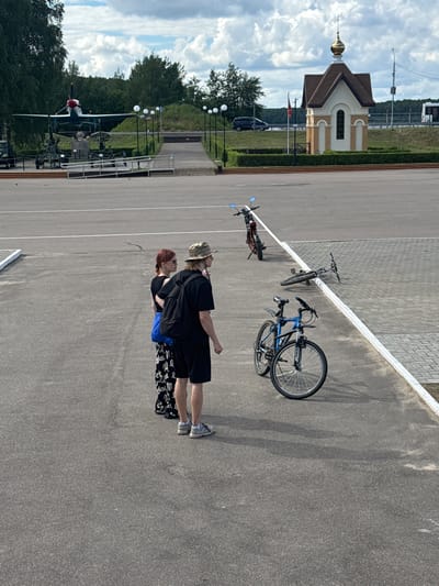 In Desnogorsk, Russia, an outdoor scene depicts two individuals standing on an asphalt surface. A woman with reddish hair, wearing a black top, blue bag, and patterned skirt, stands to the left. Beside her, a man in a camouflage hat, black t-shirt, and black shorts, wearing a backpack, faces away from the camera. In the background, a blue bicycle rests on its side, and another bicycle is upright, while a third motorcycle is parked further away. The environment features trees, a small chapel with a golden dome, and a body of water. In the distance, a historical airplane is displayed on a pedestal, part of a museum exhibit, along with military equipment. The sky is partly cloudy.