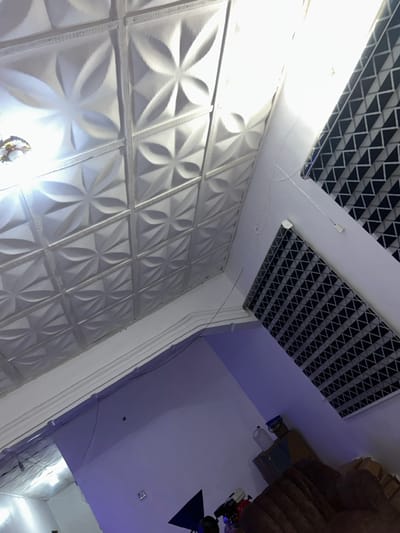 A modern interior showcases a textured white wall with repeating floral patterns, contrasted by geometric ceiling panels in black and white. A decorative wall molding accents the corner. The adjacent wall and ceiling are painted in a gradient of white to light purple. Part of a brown upholstered piece of furniture and some other objects are visible in the upper right corner of the image.