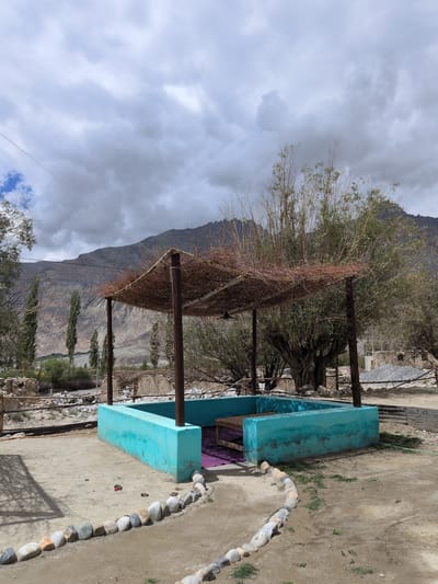 The image depicts an outdoor seating area in Panamik, India, characterized by a turquoise-colored, low-lying structure with a wooden bench inside, covered by a rustic pergola made of branches and twigs. The scene is set against a backdrop of arid, mountainous terrain with scattered trees and a cloudy sky. The ground is sandy and dusty, with a border of smooth, rounded stones lining a pathway. The overall impression is one of a simple, weathered structure in a remote, natural setting.