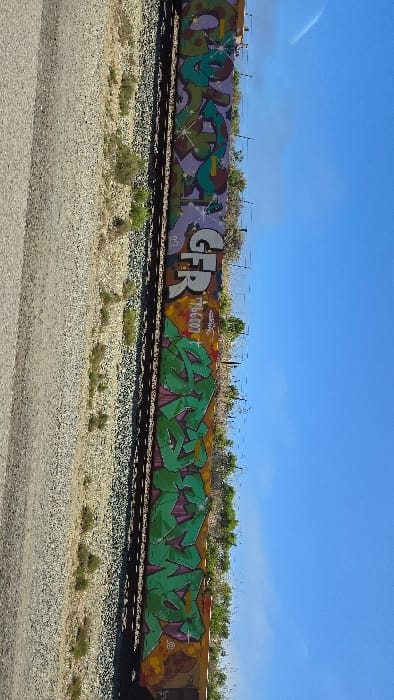 A train car showcases vibrant graffiti art. The leftmost section features purple and green designs, followed by "GFR" in bold white letters outlined in black. A section with "766607" is visible, leading to a predominantly green graffiti piece. The background includes a clear blue sky, ground cover, and railway tracks covered with gravel. The scene captures the raw, urban artistic expression on a transportation vehicle.