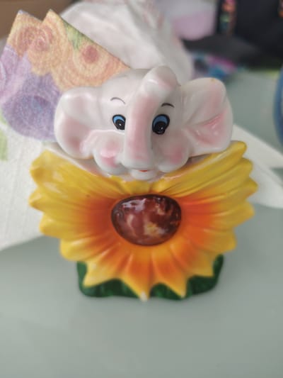 The image shows a close-up, eye-level, vertical shot of a ceramic figurine of a baby elephant emerging from a sunflower. The elephant is white with pink accents on its cheeks and ears, and has blue eyes and black eyebrows. It is positioned above a large, yellow and orange sunflower. The center of the sunflower is a dark brown, textured disc. The figurine is resting on a light grey, reflective surface, likely a table. In the background, there are blurred elements including what appears to be a white tissue and a piece of fabric with a floral pattern in shades of purple, pink, and orange. The overall lighting is bright, casting soft shadows. There is no discernible indication of the city of Aytos, Bulgaria within the image itself.