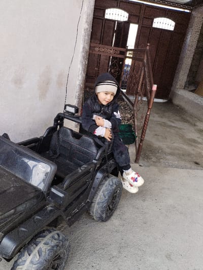A young child is seated in a black electric ride-on toy car in an outdoor paved area in the city of Limbur, Uzbekistan. The child, appearing to be a toddler, wears a light-colored beanie with a dark stripe, a dark hooded jacket with a white logo, dark pants, and white Crocs-style clogs with colorful embellishments. Their hands are clasped in front, and they are looking towards the camera with a slight smile. The black toy car resembles an off-road utility vehicle, featuring knobby tires and a forward-facing light bar mounted on a roll-bar-like structure, and appears somewhat dirty. To the left of the child, a light-colored wall shows staining. In the background, large, dark brown double doors or gates with decorative metalwork and arched upper windows are partially open. A brown, possibly wooden, upright structure stands between the child and the gates. The ground beneath is a rough concrete or paved surface, and a dark green patterned textile is visible behind the toy car.