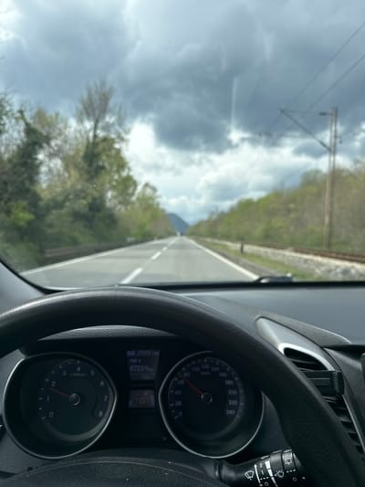 The image depicts a first-person perspective from within a moving vehicle, located in the city of Bistrice, Montenegro. The lower section of the frame is occupied by a black, textured steering wheel. Behind it, the vehicle's instrument cluster displays a tachometer on the left, with its needle indicating approximately 1100 RPM, and a speedometer on the right, showing a speed of approximately 90 km/h. A central digital display reveals an odometer reading of 259651 km, a trip meter indicating 4723.9 km, and an ambient temperature of 16°C. A black control stalk for windshield wipers is visible to the right of the steering wheel, next to a partially visible silver-trimmed air vent that contains a black phone holder.
Through the vehicle's windshield, a two-lane asphalt road is visible, extending into the distance under an overcast sky filled with dark grey clouds. The road features a solid white line separating opposing directions of traffic and dashed white lines marking individual lanes. To the left of the road, a metal guardrail runs parallel, backed by a dense array of green and light-green deciduous trees and foliage. To the right of the road, a single set of railway tracks is present on a gravel bed, also flanked by similar deciduous trees and foliage. A utility pole, likely supporting overhead lines, is discernible further down the railway. Distant hills or mountains are faintly visible on the horizon, partially obscured by the cloud cover. The vehicle is in motion, indicated by the speedometer and tachometer readings. No individuals are explicitly visible.