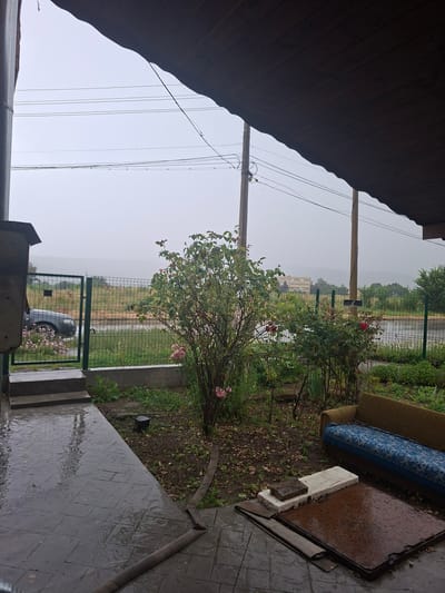 This media file captures an outdoor scene in Yasen, Bulgaria, during a rainy day. The foreground features a covered porch with a wooden ceiling, and to the right, a paved area glistening with rain. Several objects are scattered on the ground, including what appears to be an old couch with a blue and white patterned cushion, a rectangular object with a brown surface that is wet, and some discarded building materials.
Beyond the porch, a garden area is visible with rose bushes in bloom, their pink and red flowers adding a touch of color to the overcast day. A metal fence separates the garden from a grassy field with a body of water, possibly a pond or lake, in the distance. A light-colored building with multiple windows can be seen across the field, and beyond that, a hazy landscape stretches towards the horizon. A silver car is parked behind the fence, partially obscured by the vegetation.
The weather is clearly rainy, as evidenced by the wet surfaces and overcast sky. The overall mood is somber and perhaps a little melancholic, due to the rain and the presence of discarded items. There is no visible text in the image. The time of day is difficult to determine precisely due to the overcast conditions, but it appears to be daytime. The setting suggests a rural or semi-rural environment.