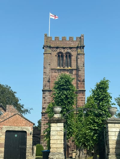 This media file captures a close-up view of a tall, ancient stone church tower bathed in bright sunlight, suggesting it's midday. The tower is constructed from reddish-brown stone with crenellated battlements at the top, indicative of medieval architecture. Intricate stonework adorns the tower, including detailed window surrounds featuring pointed arches and tracery. A St. George's flag, white with a red cross, flies proudly from a flagpole attached to the tower, hinting at a location in England.
Lush green trees with dense foliage partially obscure the tower, their leaves casting dappled shadows. They suggest a setting with well-maintained grounds, possibly a churchyard or garden. In the foreground, a section of a red brick building with a dark wooden door is visible, adding to the sense of an established, historic location. Decorative stone urns and a wrought-iron fence are also present, contributing to the ornate character of the scene. The sky is a clear, vibrant blue, indicating fair weather. The overall impression is one of historic grandeur and tranquility. The scene appears to be located in Chester, United Kingdom, given the architectural style and the flag. There is no discernible activity or event taking place; it's a still shot of a historical architectural feature.