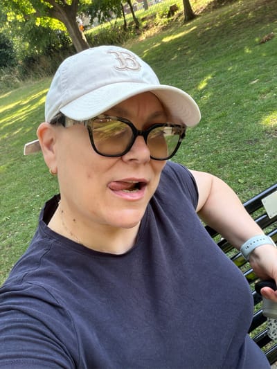 A person wearing a white baseball cap and dark t-shirt is sitting on a park bench. The person is also wearing glasses with a tortoiseshell pattern and a light blue fitness tracker on their left wrist. They are looking upwards with their mouth slightly open and tongue visible. The scene is outdoors with a grassy area and trees in the background, suggesting a park or garden setting. Sunlight filters through the trees, casting dappled shadows on the grass, indicating it is daytime with fair weather. The individual appears relaxed and engaged in an activity, possibly enjoying the outdoors. There are no explicit signs of a specific event taking place, but the setting suggests a leisurely moment. No discernible text is visible in the image, apart from a small logo on the baseball cap and some faint lettering on the bench.
