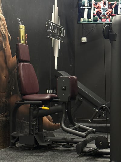 A weight training machine with padded burgundy seats is positioned against a black wall adorned with a white "AZGARD NUTRITION" logo. A television displays a bodybuilder in action. A black weight training machine is visible in the background. Part of an advertisement is also visible at the bottom of the image. The floor is carpeted in gray.