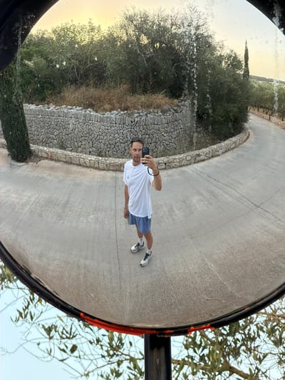 A man is taking a selfie in a convex mirror. He is wearing a white t-shirt, blue shorts, white socks, and athletic shoes. He is standing on a paved road that curves to the right. Behind him, there is a stone wall and dense vegetation. A cypress tree is visible to the left of the wall. The sky is bright, suggesting it is daytime. The scene is set in the city of Montuïri, Spain.