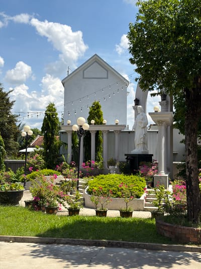 The image depicts a serene outdoor scene in Hội Tín, Vietnam. A white building with a triangular roof is visible in the background, adorned with a string of lights. In front of the building, a white statue of a robed figure holding a child stands on a dark pedestal, framed by white classical columns. The foreground is filled with lush green grass, various potted plants, and vibrant pink flowers. Several tall, slender evergreen trees are strategically placed throughout the garden, adding to the tranquil atmosphere. The sky is a clear blue with scattered white clouds. The overall impression is one of a well-maintained garden or religious sanctuary.