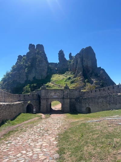 The image shows the Belogradchik Fortress, a historical monument located in the city of Belogradchik, Bulgaria. The fortress is built into and around a group of impressive sandstone rock formations. In the foreground, a cobblestone path leads towards a large arched gateway, which is part of the fortress wall. The wall itself is constructed from large stone blocks. To the left of the main gateway, there is another smaller arched opening. Behind the fortress walls, the striking rock formations rise steeply, covered in sparse vegetation. A path with railings and steps winds its way up the side of the rocks, suggesting a route for visitors. The sky is a clear, bright blue. There are no people clearly visible in the image, though the presence of paths and railings implies public access. The scene is well-lit by the sun, indicating a clear day.