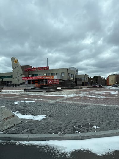 The image depicts an outdoor scene in Beloyarsky, Russia. The sky is overcast with heavy clouds. A modern building with a banner reading "НИКТО НЕ ЗАБЫТ, НИЧТО НЕ ЗАБЫТО..." (meaning "No one is forgotten, nothing is forgotten...") is prominent in the middle ground. The banner appears to be related to Victory Day celebrations. To the left, there's a concrete monument with a clock. The foreground consists of a paved pedestrian area, with some patches of snow and ice visible. In the background, there are more buildings and trees. There are no people visible in the frame.