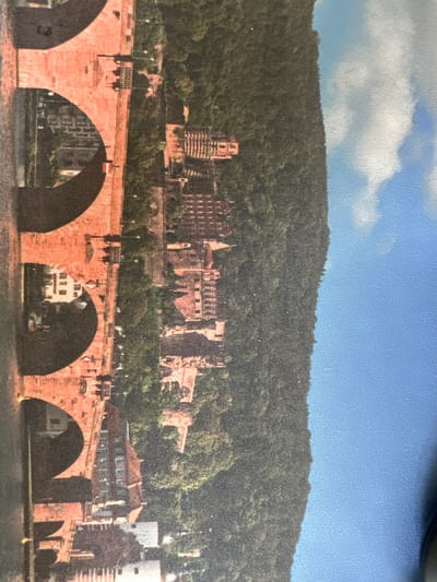 The image shows a panoramic view of Heidelberg, Germany, featuring the Old Bridge (Alte Brücke) in the foreground and Heidelberg Castle overlooking the city on the hill. The Old Bridge is composed of red sandstone arches spanning the Neckar River. The castle, also constructed of red sandstone, stands partially in ruins against a backdrop of a heavily forested hill. The sky is blue with scattered clouds. In the mid-ground are buildings lining the bank of the river. There are no people visible in the image. While Heidelberg is near Walldorf, the image is of the city of Heidelberg itself.