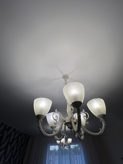 This is a low-angle, indoor photograph of an ornate, multi-armed chandelier with frosted glass lamp shades. The chandelier is made of a dark, metallic material with decorative scrollwork. Each shade has a swirling, circular pattern etched into it and is illuminated by a warm light. The chandelier is suspended from a white ceiling, which transitions to a darker wall in the background. To the right of the chandelier, a window with blinds is visible, and a patterned carpet is on the floor. The lighting suggests it could be evening or a dimly lit room. There is no visible text in the image. The scene appears to be a residential interior, possibly a dining room or living area, in Ленинское сельское поселение, Ukraine.