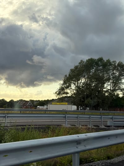 The image shows a view from a vehicle traveling on a highway in Väröbacka, Sweden. In the mid-ground, there is a building with a yellow sign that reads "Idala Trappan". Adjacent to this building, a large deciduous tree with full foliage stands prominently. To the left of the building, a red-roofed house is visible, along with a white truck and a light-colored trailer. The sky is dramatic with a mix of dark, heavy clouds and brighter, lighter patches, suggesting overcast conditions with some sunlight breaking through. The foreground features a metal guardrail and grassy vegetation.