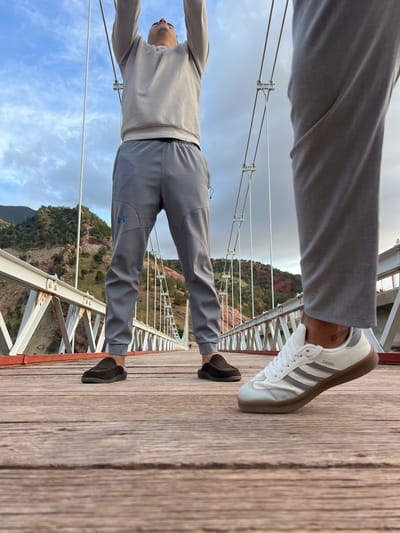 A man stretches on a suspension bridge. He wears gray athletic wear and different shoes on each foot: a white Adidas Samba on his right and a dark-colored loafer on his left. The bridge has a wooden plank surface and white and red metal support structures. Below, a mountainous landscape is visible. The sky is partly cloudy. The location is near Tabaroucht, Morocco, as evidenced by the landscape.