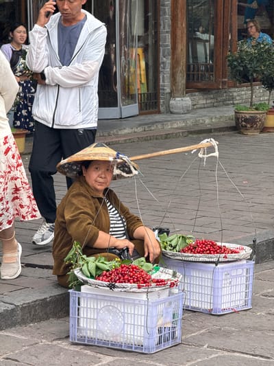 A woman wearing a conical hat, a brown corduroy jacket over a striped shirt, and athletic shoes, sits on a cobblestone surface next to two lavender plastic crates filled with red cherries and green leaves. The cherries are on platters within the crates. The woman's carrying device is made of a wooden stick attached to wire frames that hold the crates. A man in a white jacket and black pants looks at his cell phone, partially visible to the left of the woman. Parts of two other people are visible, with one wearing a dress with a red floral pattern. The background shows a building with windows and a plant in a pot. The scene is likely in Fenghuang, China.