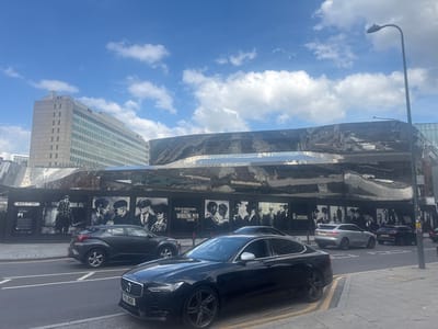 The image depicts a street scene in Birmingham, United Kingdom, likely near Grand Central station, identifiable by a building with "GRAND CENTRAL" signage in the background. A modern, undulating building with a highly reflective metallic facade dominates the middle ground, adorned with a series of large black and white murals. These murals appear to be promotional or artistic displays, featuring portraits of men in period attire, some with quotes, evoking a historical or cinematic theme.
In the foreground, a black Volvo S90 sedan is parked on the right side of the road, its license plate "FVL 810X" partially visible. To the left of the Volvo, a dark grey hatchback, possibly a Toyota C-HR, is in motion on the street, heading away from the viewer. Further down the street, a silver Jaguar F-Pace is seen turning, and a black Land Rover Discovery is parked. The street is paved with asphalt and bordered by a paved sidewalk. A tall, modern street lamp stands on the right side of the image, its light fixture pointing towards the sky. The sky is a bright blue with scattered white clouds. The overall environment suggests an urban setting during daylight hours.