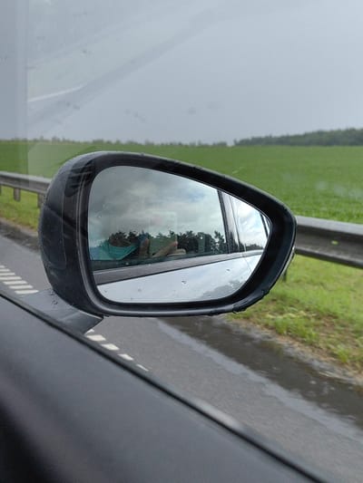 The image shows a side-view mirror of a car, reflecting a person holding a smartphone and wearing a turquoise face mask. The background in the mirror shows trees. Outside the mirror, the car is driving on a wet road next to a grassy field and a guardrail. The sky is overcast and appears to be raining. The location is identified as Дзяржынскі сельскі Савет, Belarus.