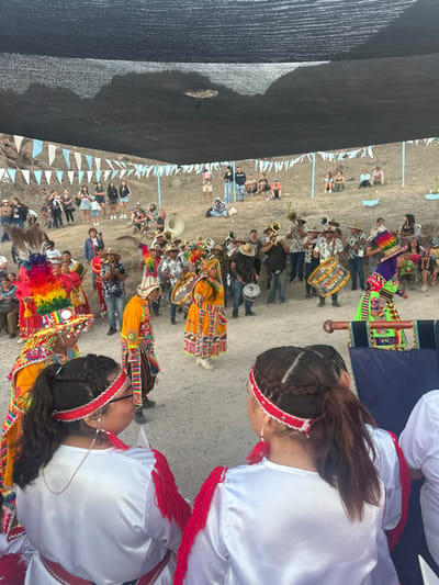 A vibrant parade unfolds outdoors. Two women in white dresses with red trim stand in the foreground. Behind them, people in colorful traditional costumes march, accompanied by a brass band. Many onlookers watch from the sides. White pennant flags line the route. A large black shade structure covers part of the scene. The event appears to be a cultural celebration or festival.