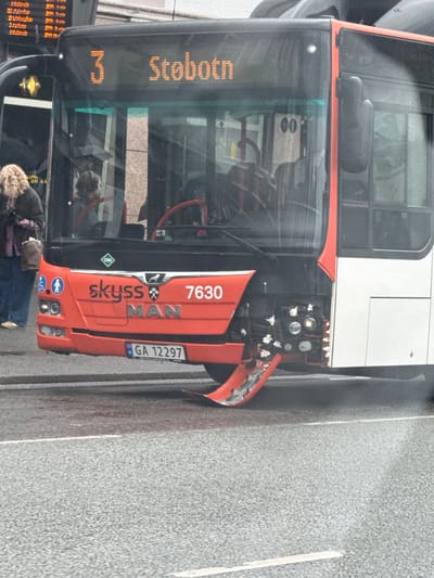 The image shows the front of a damaged red and white Skyss bus with the number "3" and "Stobøtn" displayed on its digital sign. The bus has a damaged front bumper and what appears to be some type of repair or protective material affixed to the lower front section, likely a result of a collision or road debris. The license plate reads "GA 12297". The bus is parked on a street with a wet asphalt surface, indicating recent rain. To the left of the bus, a person with curly blonde hair is standing, holding a dark handbag and looking towards the bus. Behind the person, part of a building with windows and a sign is visible. The environment suggests an urban setting in Bergen, Norway, given the "Skyss" branding, which is the public transportation company for Vestland county.