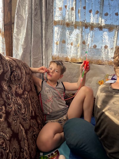 A young boy with blond hair and blue eyes lies on a brown patterned couch, holding a colorful toy connected to a red tube. He is wearing a grey t-shirt with red and black trim and patterned shorts. The boy looks towards the camera with a neutral expression. Sunlight streams through a sheer curtain with floral embroidery behind him, suggesting it's daytime. The room appears to be a living space, possibly in a home, with the couch as the central element. Another person's arm and shoulder are visible at the top of the frame, partially obscured. There is a glimpse of a blue blanket and a product package with Cyrillic text near the boy's legs. The overall scene is casual and domestic.