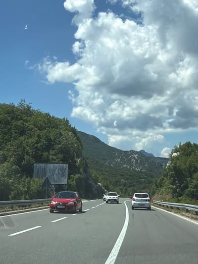 A red car is driving on a highway in Obzovica, Montenegro. The car is in the left lane and is moving away from the camera. The highway is surrounded by lush green trees and mountains. The sky is blue with some white clouds. There are other cars on the highway as well. A white car is in front of the red car, and another white car is in the right lane. The red car's license plate is visible and appears to be from Montenegro. The scene is lit by daylight.